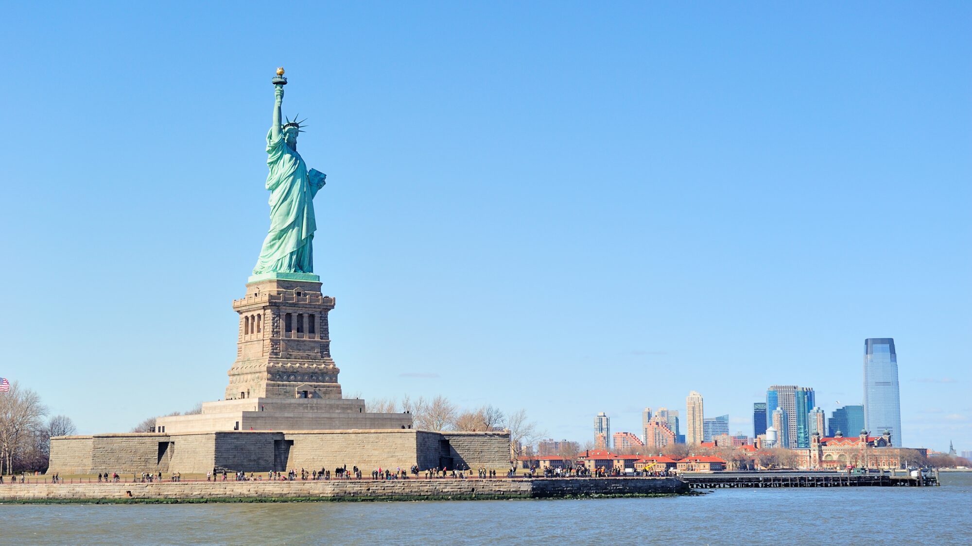 Statue of Liberty faces New York City Manhattan downtown skyline with skyscrapers over Hudson River with clear blue sky.